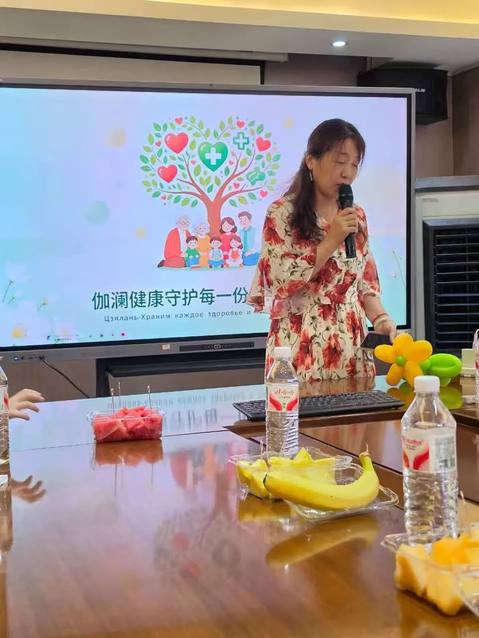 Speaking into a microphone in a conference room in front of a presentation screen, a table with fruit and water in the foreground