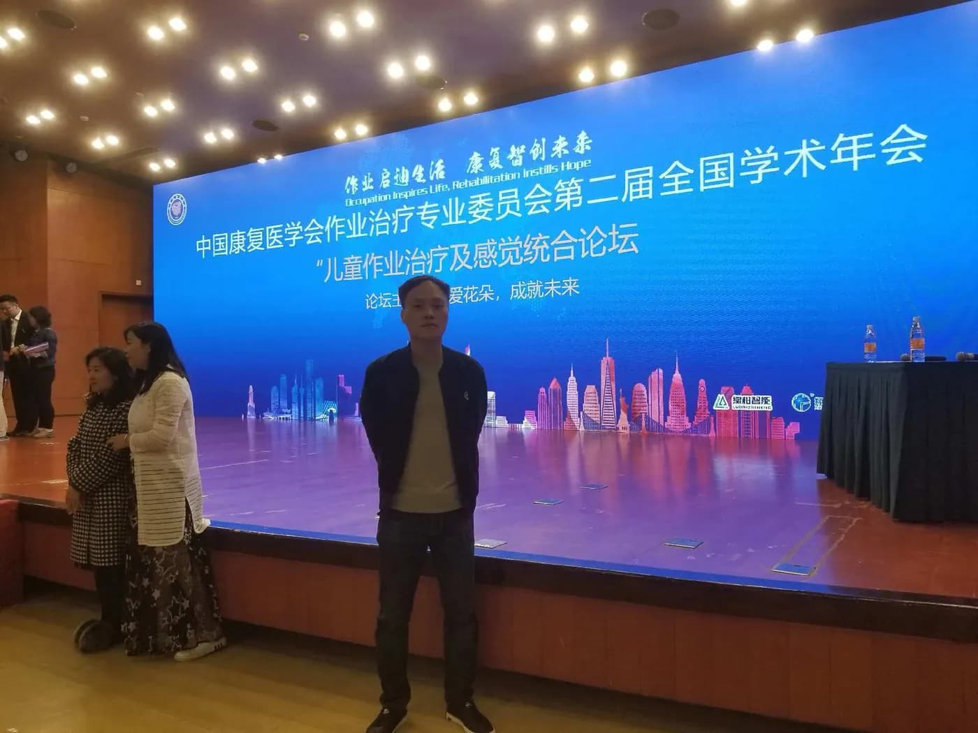 一名男子在会议厅舞台前，身后屏幕显示儿童作业治疗及感觉统合论坛的名称